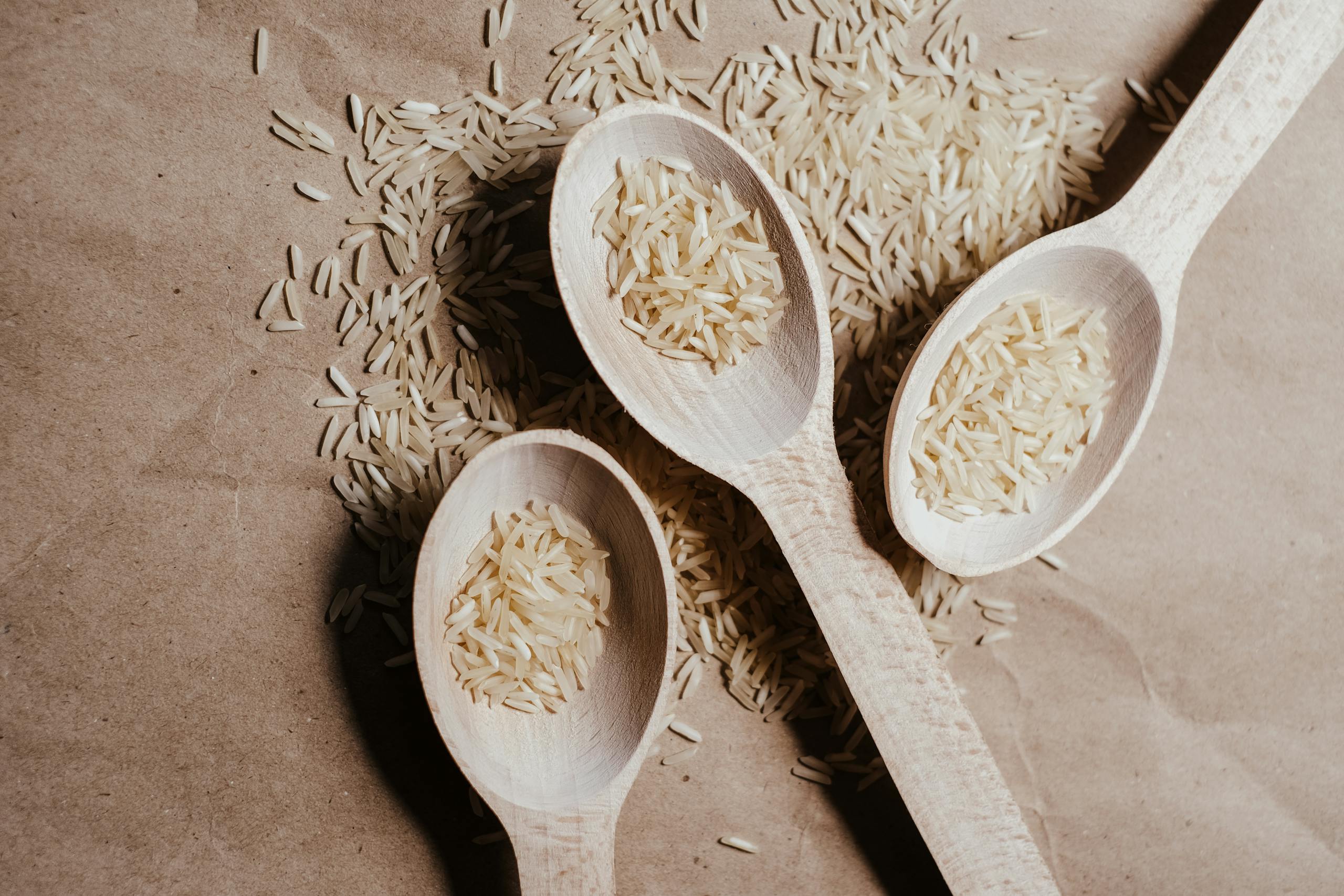 Photo by Vie Studio Three wooden spoons filled with uncooked rice on a textured brown paper surface.